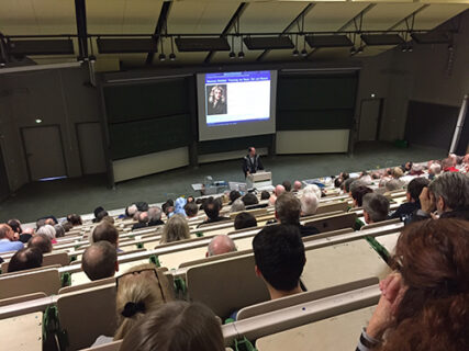 Das Foto zeigt den vollbesetzten Hörsaal G bei der Vorlesung Moderne Physik am Samstagmorgen. Prof. Mecke hält eine Präsentation und steht am Rednerpult, hinter ihm auf der Leinwand sieht man ein Foto von Newton.