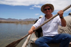 Das Foto zeigt Prof. Daesslé-Heuser in einem Kanu sitzend auf dem Colorado River. Er trägt ein weißes Hemd, Bluejeans und einen Sonnenhut. Er hat ein Paddel in der Hand, eine Sonnenbrille auf und lächelt.