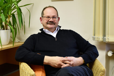 Portrait Prof. Dr. Jürgen Schatz, der in seinem Büro auf einem Stuhl sitzt und die Hände vor sich verschränkt hat. Im Hintergrund sieht man eine Grünpflanze im Topf auf dem Fensterbrett stehen.