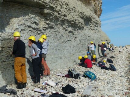 Studierende stehen an einer Felswand. Sie tragen alle gelbe Schutzhelme und betrachten die Gesteinsformation.
