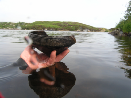 Das Foto zeigt den See Loch Arnish. Im Hintergrund sieht man das grüne Ufer. Eine Hand taucht aus dem Wasser hervor und hält eine Gefäß, eine Unstan Bowl, in die Kamera. Die Unstan Bowl ist rund und aus dunklem Material, das aussieht wie Stein. Es läuft unten spitz zu und hat oben einen mit drei Rillen verzierten Rand. Auf einer Seite ist das Gefäß beschädigt.