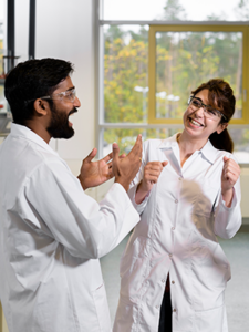 Auf dem Foto sieht man zwei Menschen in einem Chemie-Labor. Die junge Frau und der junge Mann tragen beide Laborkittel. Beide sehen glücklich aus und haben die Hände erhoben, weil sie sich freuen. Der Mann hat schwarze Haare und einen schwarzen Vollbart und lacht. Die Frau rechts neben ihm blickt ihn an, lacht ebenfalls und hat langes schwarzes Haar.