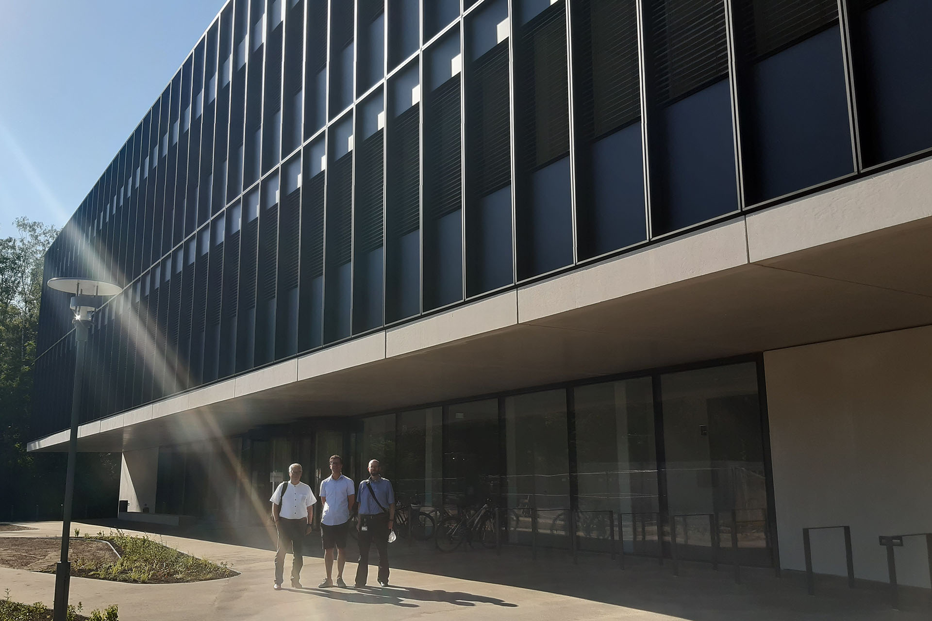 Das Foto zeigt das neue ECAP-Gebäude. Es scheint die Sonne und der Himmel ist blau. Vor dem Gebäude stehen drei Männer in sommerlicher Kleidung, die in die Kamera blicken. Links steht Kanzler Zens, in der Mitte Prof. Dr. Stefan Funk und rechts ein weiterer Mann.