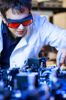 Das Foto zeigt einen jungen Wissenschaftler mit kurzen dunklen Haaren. Er trägt eine Laserschutzbrille und einen Laborkittel über einem dunkelblauen Hemd. Er steht in einem Laserlabor vor eineme Laser und berührt mit einer Hand die Gerätschaften. Er sieht konzentriert aus.
