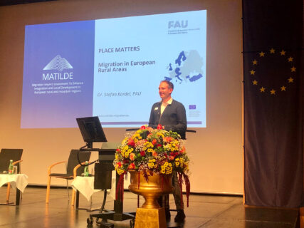 Dr. Stefan Kordel vom Institut für Geographie der FAU präsentiert die Projektergebnisse von MATILDE. (Bild: David Spenger) Auf dem Foto steht Herr Kordel auf einem Podium und spricht ins nicht zu sehende Publikum. Er trägt ein weißes Hemd, einen dunklen Pullover und eine braune Hose. Er steht leicht verdeckt von einem Blumenbuquet und blickt auf einen Monitor, der vor ihm aufgebaut ist. Im Hintergrund sieht man eine Leinwand, auf der das Logo von MATILDE zu sehen ist sowie der Titel seines Vortrags: "Place Matters: Migration in European Rural Areas". Rechts im Bild hängt eine Europafahne von der Wand. Im Hintergrund stehen mehrere ungenutzte Stühle und ein Tisch, auf dem Getränke stehen.