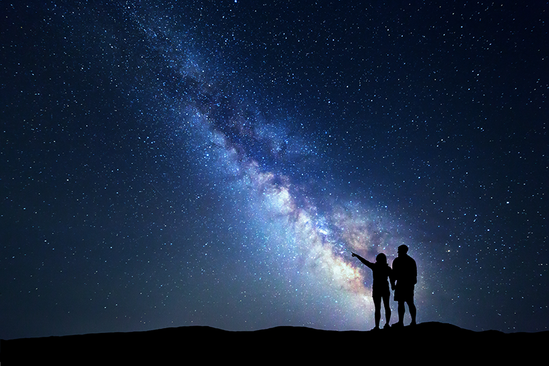 Das Foto zeigt die Milchstraße. Im Vordergrund sieht man schwarz die Silhouette eines Mannes und einer Frau. Die Frau deutet in den Sternenhimmel.
