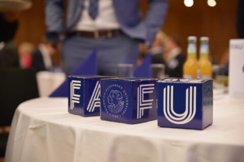 Das Foto zeigt drei Pappwürfel, auf denen FAU steht. Die Würfel stehen auf einem festlich geschmückten Tisch, im Hintergrund sieht man unscharf Gäste.